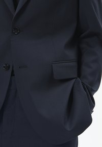 Sötétkék öltönyzakó, álló hajtókával, egygombós záródással és egy kis zsebpokkal a bal oldalon. Sima textúra és szabott illat.