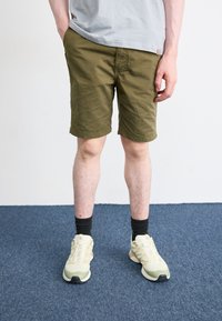 Shorts vert olive en tissu légèrement froissé, associés à des chaussures de sport claires et des chaussettes noires.