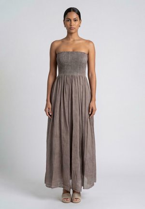 Vrouw met donker haar die een strapless, bruine, lange jurk tot op de vloer en beige open-teen schoenen draagt, staat voor een effen lichte achtergrond.