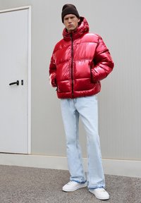 Jachetă puffer roșie cu un finisaj lucios, fermoar frontal și glugă. Asortată cu blugi albaștri deschiși și adidași albi pe o suprafață texturată.