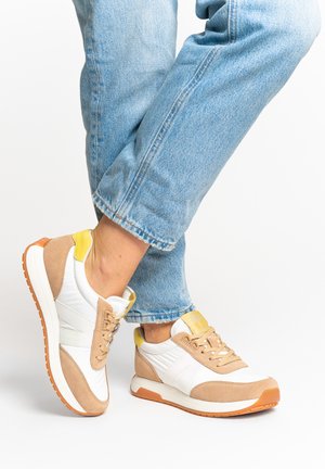 Person wearing beige, white, and yellow sneakers with gum soles, paired with rolled-up light blue denim jeans, standing on a white background.