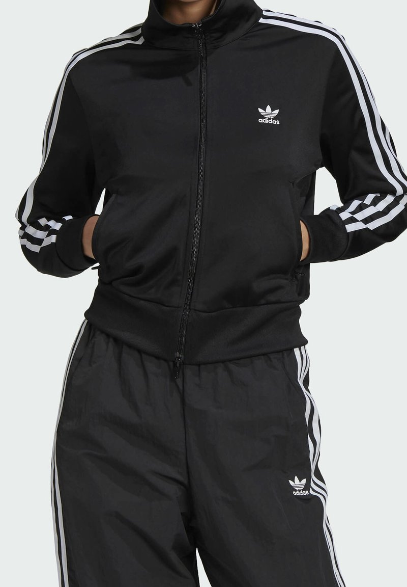 Svart zip-up träningsjacka med hög krage, vit Adidas-logga på bröstet och tre vita ränder längs ärmarna. Matchas med svart byxa i samma stil.