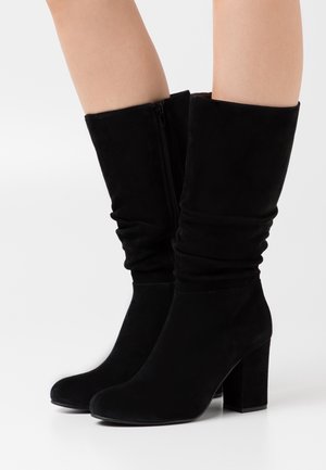 Bottes noires en daim montant jusqu'au genou avec un design décontracté, talon bloc et fermeture éclair latérale. Texture lisse avec un bout arrondi et un style minimaliste.