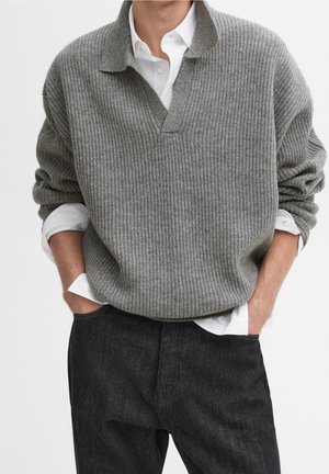 Mężczyzna w szarym prążkowanym swetrze noszonym na białej koszuli z kołnierzykiem, z podwiniętymi rękawami, w ciemnych jeansach, z rękami w kieszeniach.