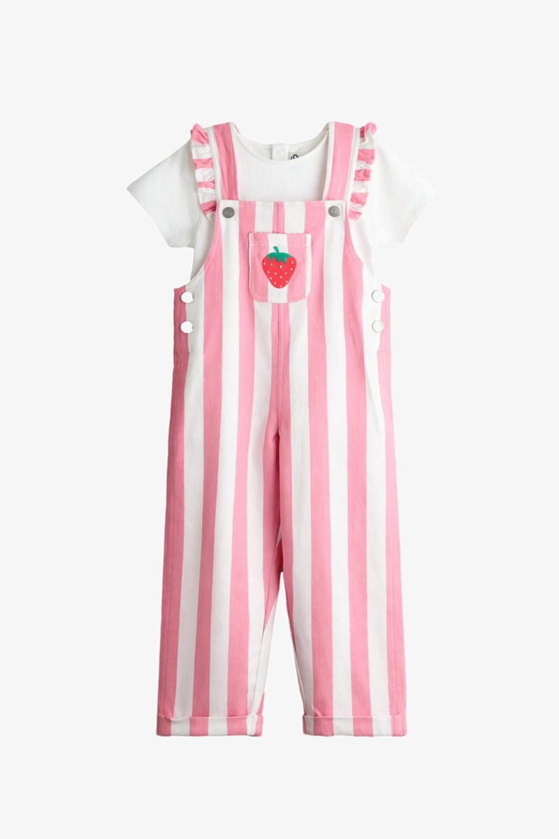 Camicia bianca a maniche corte sotto una salopette a righe verticali rosa e bianche con bretelle arricciate e una tasca frontale con disegno di una fragola rossa.