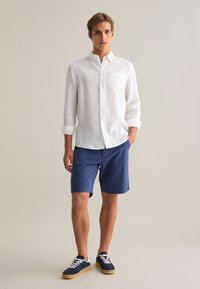 Camisa de manga larga blanca de lino con un bolsillo en el pecho, combinada con pantalones cortos texturizados en azul marino y zapatillas azul marino con cordones blancos y suelas beige.