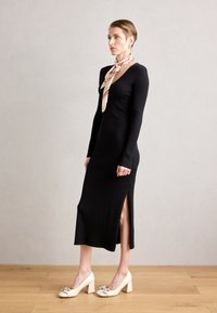 Robe midi noire côtelée avec des manches longues et une fente sur le côté, associée à des chaussures à talons blocs crème ornées d'un nœud et d'accents en argent.