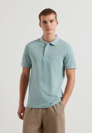 Junger Mann mit kurzen Haaren, der ein hellblaues kurzärmeliges Poloshirt und beige Hosen trägt, steht mit einer Hand in der Tasche.