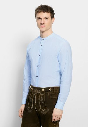 Junger Mann trägt ein hellblaues Hemd mit Stehkragen und dunkelbraune bestickte traditionelle Lederhose, steht vor einem schlichten Hintergrund.
