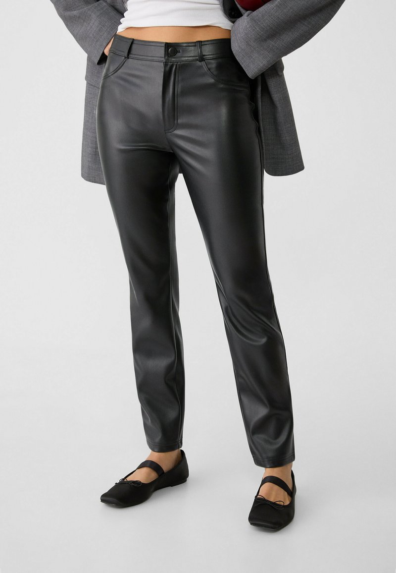 Pantalons en simili cuir noir à coupe ajustée, dotés de poches avant, d'une fermeture à bouton et d'une texture lisse et brillante. Associés à des chaussures plates noires.