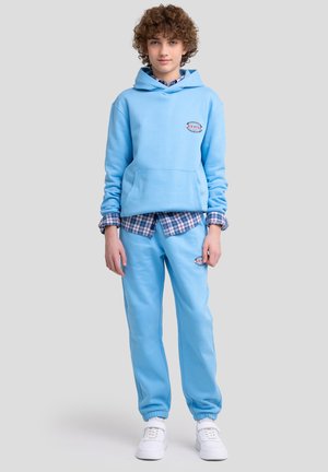 Lichtblauw sweatshirt met capuchon en voorzak, gecombineerd met bijpassende joggingbroek. Heeft een logo-label en een geruit overhemd eronder. Witte sneakers.