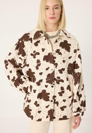 Žena s rovnými blond vlasmi, ktorá nosí nadmerne veľkú krémovú košeľu s nepravidelným hnedým vzorom kravy, stojí s jednou rukou v vrecku pred jednoliatym pozadím.