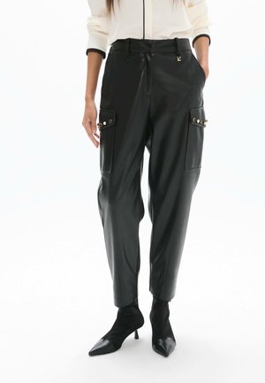 Mujer con pantalones cargo de cuero negro con bolsillos abotonados y botines negros de punta afilada, combinados con una camisa blanca.
