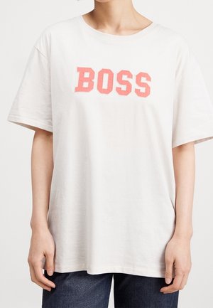 Persoon draagt een loszittend wit T-shirt met grote roze tekst "BOSS" op de borst en donkere broek, staand tegen een effen lichte achtergrond.