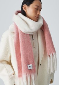 Sciarpa con gradiente rosa e bianco realizzata in morbido tessuto, con frange alle estremità e un piccolo etichetta del marchio. Indossata sopra un cappotto di lana color crema.