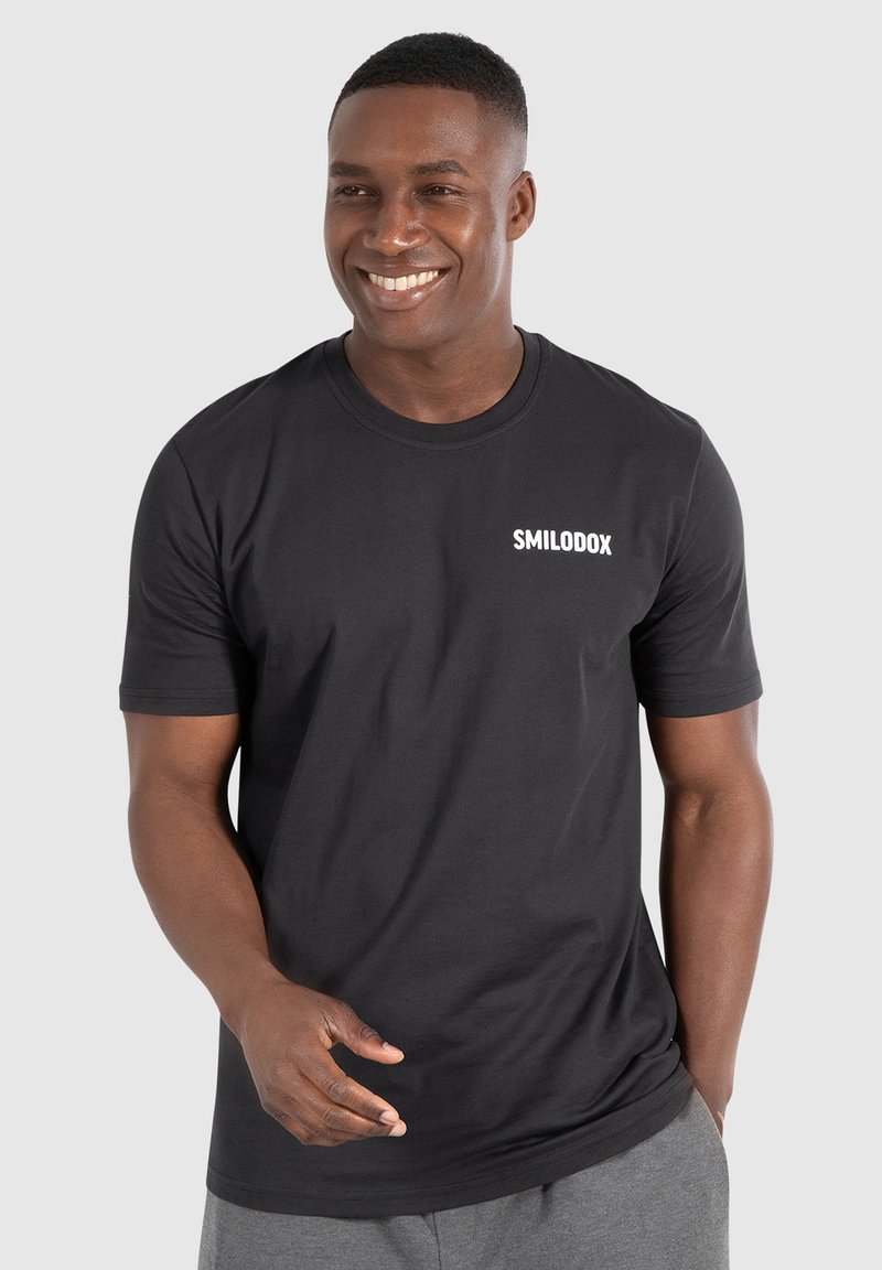 Sort bomulds T-shirt med korte ærmer og rund halsudskæring, med et hvidt "SMILODOX" logo på venstre brystområde. Glat texture.