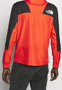 Červená a černá větrovka s kapucí, elastickými manžetami a logem na levém rameni. Hladká textura, lehký design.