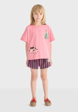 Ungt barn iført lyserød T-shirt med illustrationer af landmand og ko, stribede lilla og grønne shorts samt brune sko med røde snørebånd, stående på hvidt gulv.
