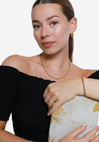 I gioielli in oro includono una collana con catena attorcigliata, un bracciale intrecciato e un anello. Il modello indossa un top nero senza spalline, tenendo un libro con motivo.