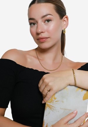 Collier en chaîne dorée, bracelet doré et bague sur une femme tenant un carnet à motifs. Haut noir à épaules dénudées, peau lisse et cheveux raides.