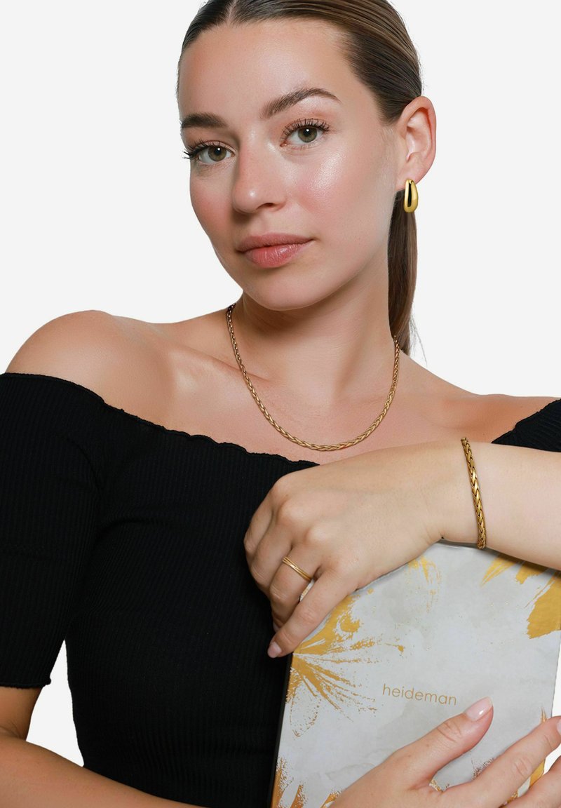 Gouden sieraden omvatten een gedraaide kettinghalsketting, een gevlochten armband en een ring. Het model draagt een zwart topje zonder schouders en houdt een patroonboek vast.