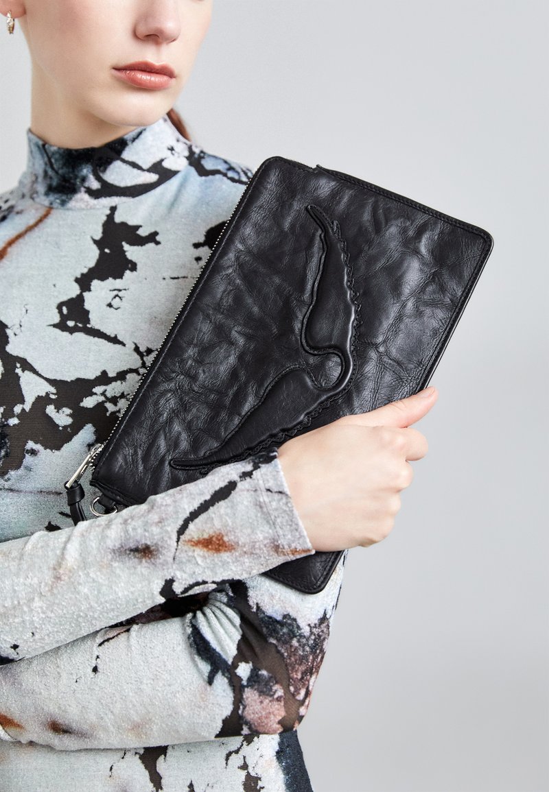 Femme tenant une grande pochette en cuir noir avec un motif abstrait embossé, portant un haut à col haut, manches longues et motif.