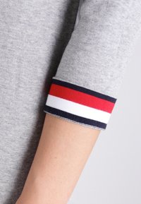 Pullover grigio con una vista ravvicinata della manica che mostra un polsino a righe in rosso, bianco e blu navy. Tessuto morbido, design informale.