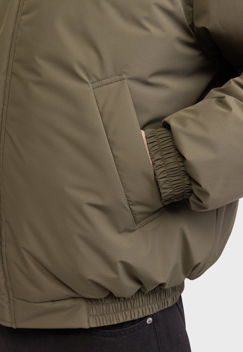 Giacca imbottita verde oliva con una finitura texturizzata, polsini elasticizzati e tasca laterale. Progettata con un orlo arricciato e dettagli di cucitura minimi.