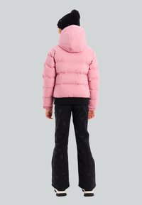 Persona con una chaqueta acolchada rosa con capucha, pantalones negros con estampado de estrellas, zapatos blancos y un gorro negro de punto, de espaldas sobre un fondo gris.