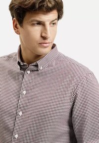 Mężczyzna w szaro-bordowej koszuli w kratę z guzikami, patrzący w lewo na tle jednolitego jasnego tła.