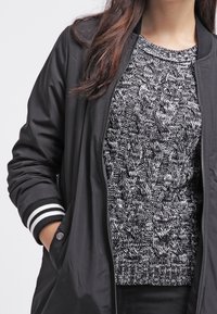 Giacca bomber nera, leggera, con polsini a righe, indossata sopra un maglione a coste in bianco e nero. Visione ravvicinata.