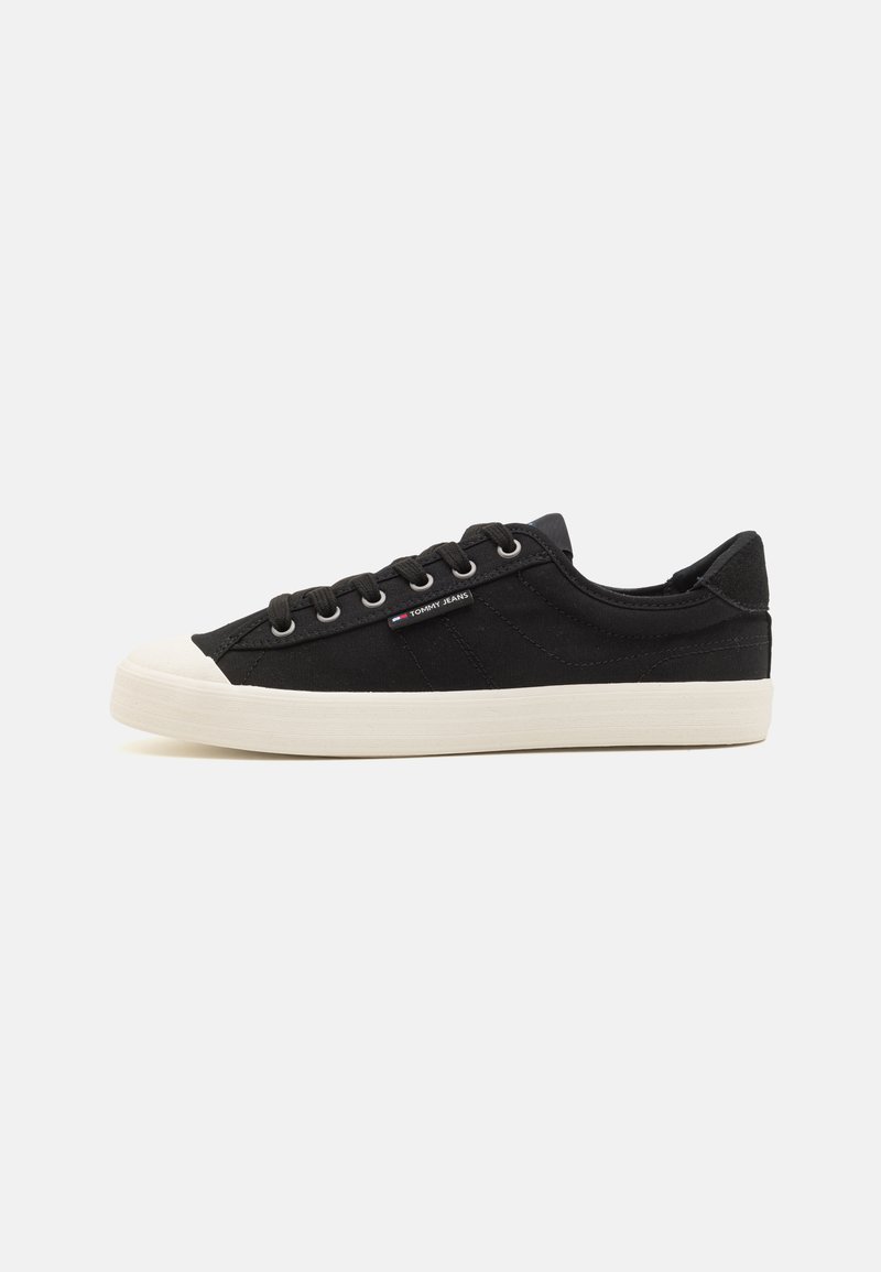Svart kanvas sneakers med hvit gummitå og såle, med seks øyestrikk, svarte snøringer og en liten logo aksent på siden.