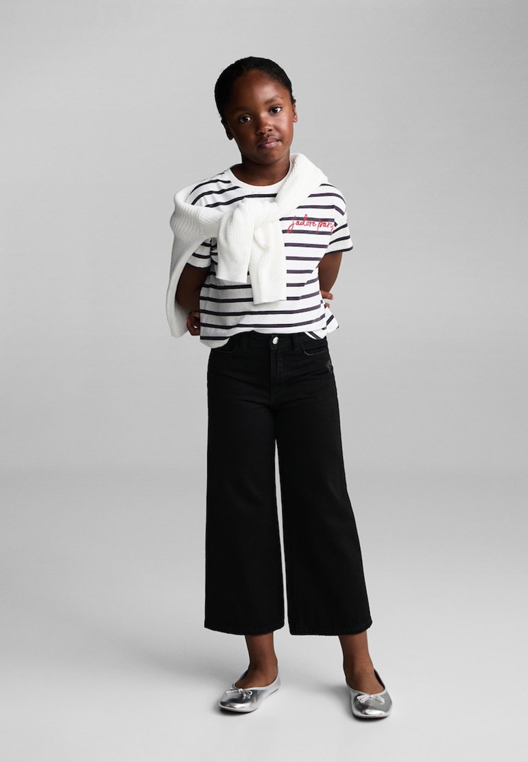 Enfant portant une chemise à rayures à manches courtes avec des lettres rouges, un pantalon large noir, et des chaussures plates argentées, avec un pullover blanc drapé autour des épaules.