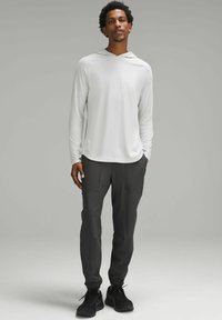 Hellgraues langärmliges Kapuzenshirt kombiniert mit dunkelgrauen schmal zulaufenden Hosen. Die Hose hat zwei Seitentaschen und elastische Bündchen. Schwarze Sportschuhe.