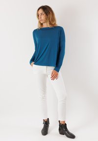 Maglietta a maniche lunghe di colore teal in tessuto leggero, abbinata a pantaloni bianchi slim-fit e stivaletti neri con cerniere. Design semplice e moderno.