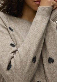 Beżowy sweter wykonany z miękkiego materiału, z czarnymi wzorami w kształcie serca, prążkowanym dekoltem i wykończeniem mankietów, prezentujący zbliżenie na teksturowany materiał.