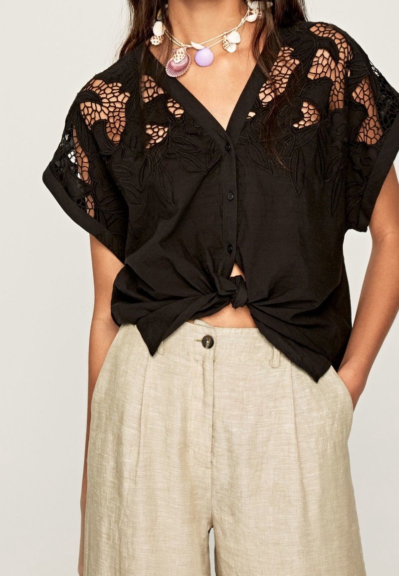 Haut noir à manches courtes avec boutons et épaules en dentelle florale, noué à la taille, associé à un pantalon en lin beige taille haute.