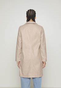 Osoba s pletenými vlasy, oblečená v dlouhém kabátu světle béžové barvy a světlých modrých džínách, stojící čelem k hladké bílé zdi.