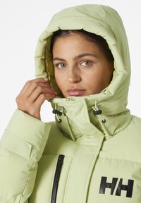 Giacca isolante verde chiaro con colletto alto e cappuccio, dotata di tasca con zip nera e logo sul petto. Tessuto testurizzato e imbottito.