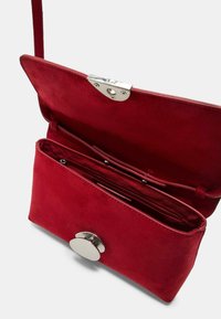 Červená semišová kabelka cez rameno so strieborným okrúhlym zapínaním, vybavená priestranným vnútrom s viacerými priehradkami a hladkým povrchom.
