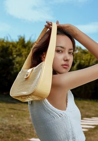 Borsa a spalla gialla con effetto crocodile, dalla forma strutturata, con pattina frontale piatta e chiusura dorata. Il modello la tiene contro uno sfondo verde sfocato.