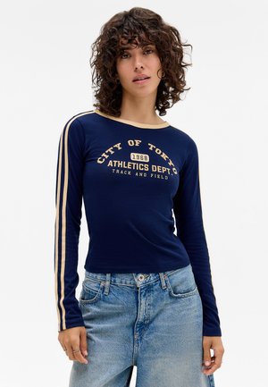 Haut à manches longues bleu marine avec un bord crème contrastant et un texte graphique "VILLE DE TOKYO 1968 DÉPARTEMENT D'ATHLÉTISME", associé à un jean bleu clair.