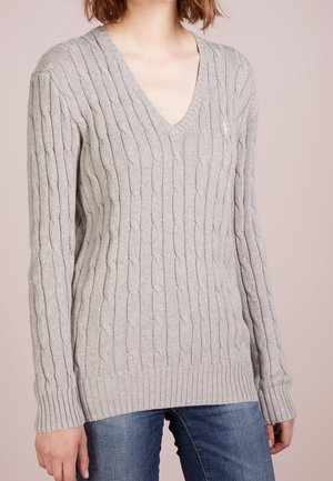 Woman wearing a light gray cable-knit V-neck sweater with long sleeves and blue jeans, standing against a plain background.