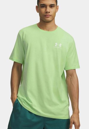 Jonge man die een lichtgroen Under Armour T-shirt en donkergroene sportshort draagt, staand met één hand in zijn zak.