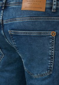 Niebieskie dzinsy z dzianiny z skórzaną metką z napisem "Langerto Knit Denim", pomarańczowymi przeszyciami i guzikiem na tylnej kieszeni.