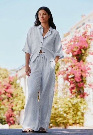 Jeune femme portant une chemise ample gris clair à boutons et un pantalon large, debout à l'extérieur avec des fleurs roses et de la verdure en arrière-plan.