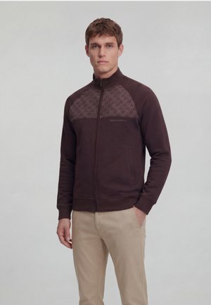 Young man wearing a dark brown zip-up jacket with patterned upper panel and beige pants, standing against a plain light background.