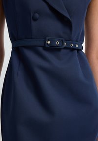 Gros plan d'une robe bleu marine avec une ceinture assortie comportant des œillets en argent et une boucle rectangulaire recouverte de tissu.