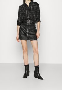 Schwarz-weiß kariertes Hemd mit Kragen, kombiniert mit einem schwarzen Leder-Minirock mit Frontreißverschluss. Schwarze spitze Stiefeletten vervollständigen den Look.