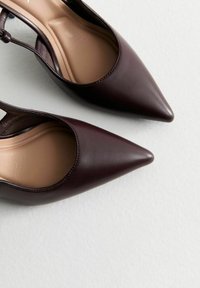 Scarpe con tacco alto di un oscuro borgogna, con punta affusolata, esterno in pelle liscia e stile aperto sul retro. Suola interna di colore chiaro visibile.